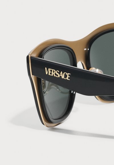 Versace sunglasses featuring black and gold frames, gray lenses, with gold logo detail on the temple. Smooth texture, oversized design.