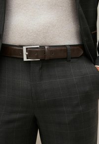 Pantalons de costume à carreaux foncés avec un motif de grille subtil, associés à une ceinture en cuir marron avec une boucle rectangulaire en argent et un pull beige clair.