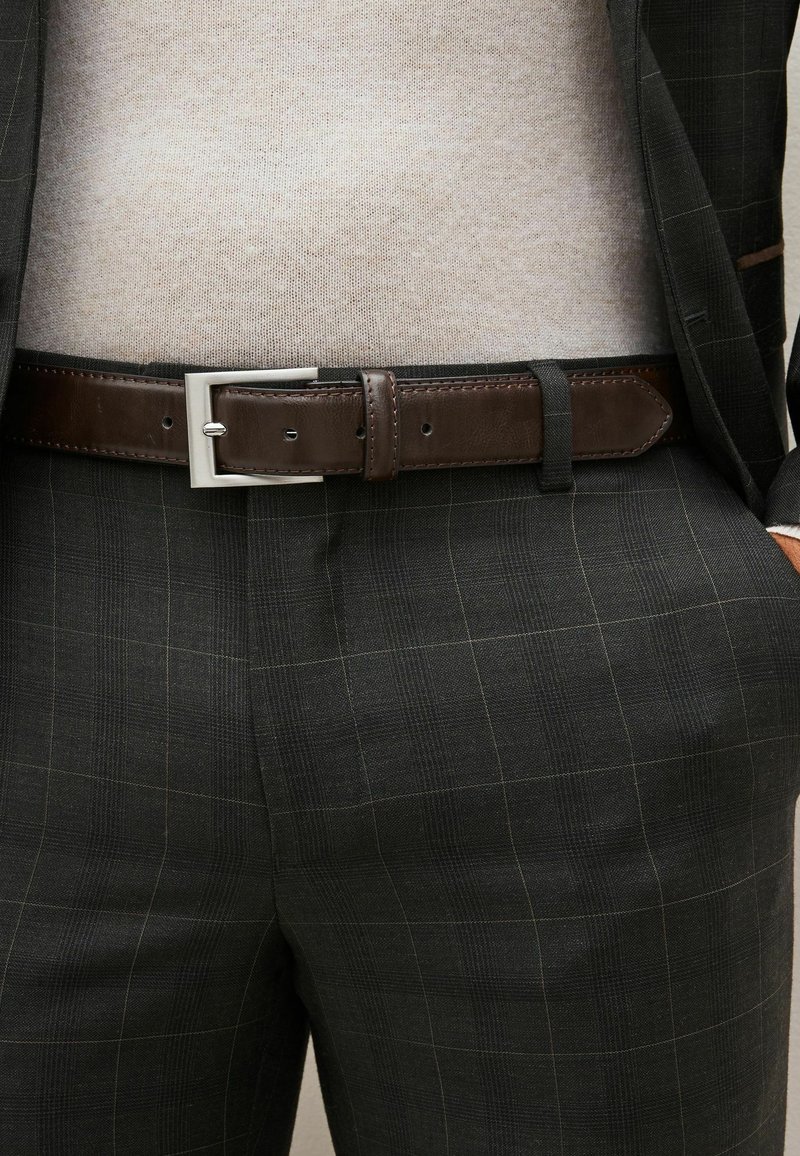 Pantalons de costume à carreaux foncés avec un motif de grille subtil, associés à une ceinture en cuir marron avec une boucle rectangulaire en argent et un pull beige clair.