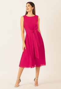 Abito plissettato fucsia con scollatura rotonda, vita con cintura e orlo fluente. Le scarpe sono sandali argentati con cinturini. Il tessuto testurizzato aggiunge profondità.