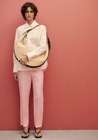 Beige hobo bag with black trim, lightweight fabric, worn on shoulder; model in white shirt, pink linen pants, and black sandals.