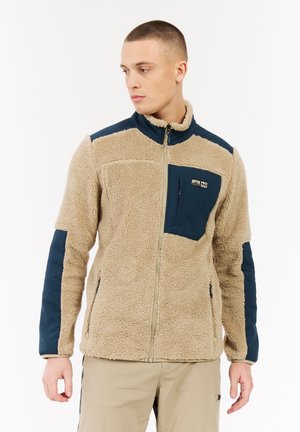 Jonge man die naar links kijkt, gekleed in een beige en marineblauwe fleecejack met borstzak en beige broek tegen een witte achtergrond.