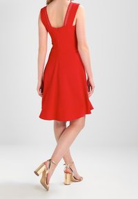 Robe rouge sans manches avec une jupe évasée, ornée de dentelle sur les bretelles. Associée à des sandales à talons dorés.