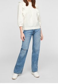 Blusa bianca con texture e maniche a volant, abbinata a jeans svasati azzurri chiari. La modella indossa sneakers bianche, in piedi su uno sfondo semplice.