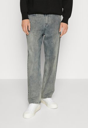 Osoba ubrana w wyblakłe szare dżinsy o prostych nogawkach, białe trampki i czarną bluzkę z długim rękawem, stojąca na tle jednolitego tła.