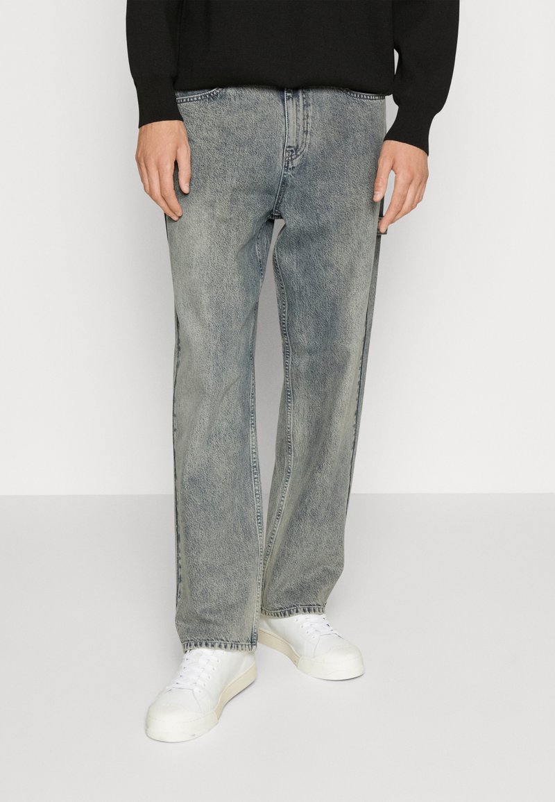 Personne portant un jean gris délavé à jambes droites, des baskets blanches et un haut noir à manches longues, debout devant un fond uni.