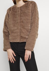 Bruine jas van imitatiebont met horizontale strepen, ronde hals, knopen aan de voorkant en lange mouwen, gecombineerd met een zwarte broek. Zachte textuur.