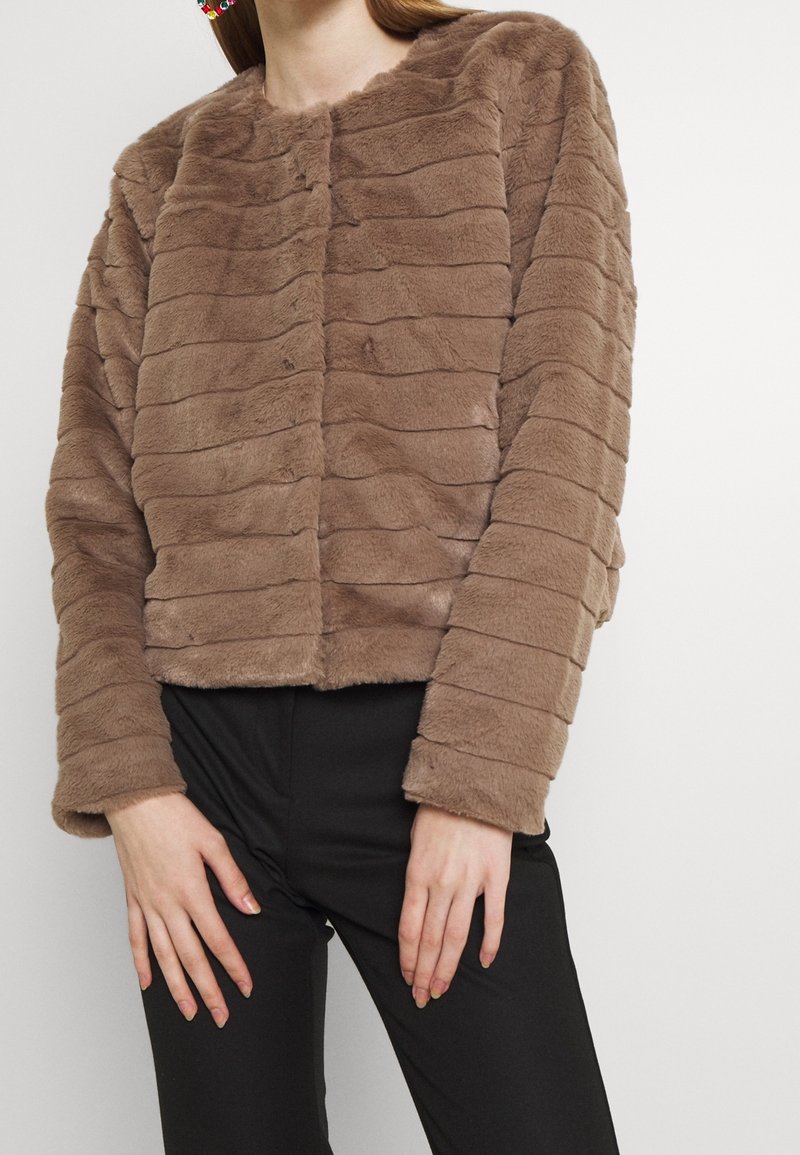 Bruine jas van imitatiebont met horizontale strepen, ronde hals, knopen aan de voorkant en lange mouwen, gecombineerd met een zwarte broek. Zachte textuur.