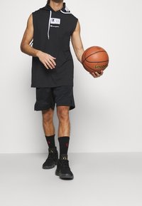 Black sleeveless hoodie with logo, paired with black shorts and socks. Holding a basketball, wearing black athletic shoes with gold accents.