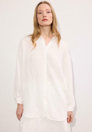 Νεαρή γυναίκα με μακριά ξανθά μαλλιά, που φοράει μία άνετη λευκή πουκαμίσα με μακριά μανίκια, πάνω σε ένα απλό ανοιχτό φόντο.