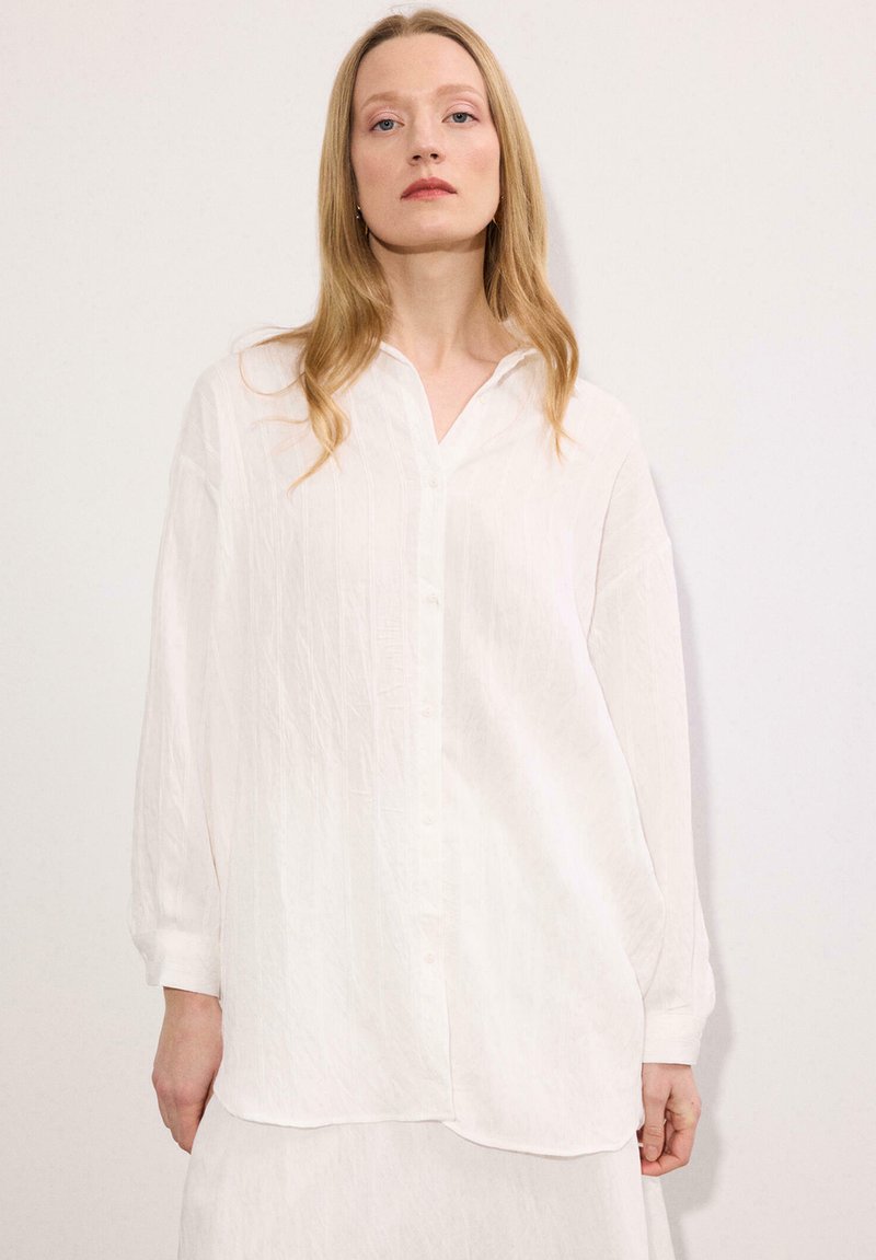 Νεαρή γυναίκα με μακριά ξανθά μαλλιά, που φοράει μία άνετη λευκή πουκαμίσα με μακριά μανίκια, πάνω σε ένα απλό ανοιχτό φόντο.