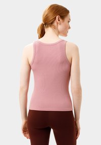 Růžový žebrovaný top na ramínka s kulatým výstřihem a úzkými ramínky, v kombinaci s hnědými legínami. Zůstává hladký a přiléhavý ve svém designu.