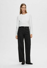 Camicia bianca a maniche lunghe, jeans neri a gamba larga con cintura, texture uniforme. Il modello è in piedi contro uno sfondo chiaro e semplice. Ballerine nere.
