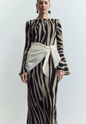 Mujer con un vestido de manga larga con estampado de cebra en negro y marrón, con un gran lazo de tela blanca atado a la cintura, de pie frente a un fondo liso.