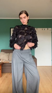 Vrouw met kort donker haar, gekleed in een doorschijnend zwart textuur topje en brede grijze broek, staande in een woonkamer met groene muren en een bruine bank.