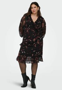 Mujer con cabello largo y oscuro, vestida con un midi negro de flores con volantes, mangas largas transparentes, medias negras y botas de tacón negro hasta el tobillo, de pie en posición recta.