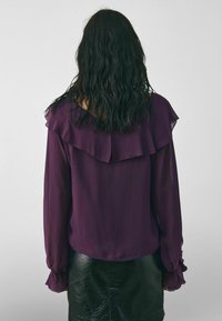 Kvinde med vådt, mørkt hår iført en lilla bluse med lange ærmer og flæser ved kraven og manchetterne, kombineret med en sort læderskirt, vendt væk fra kameraet.