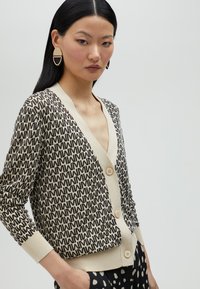 Pleteni kardigan s ponavljajućim crnim i krem uzorkom, s v-izrezom, dugim rukavima i tri krem dugmeta sprijeda.