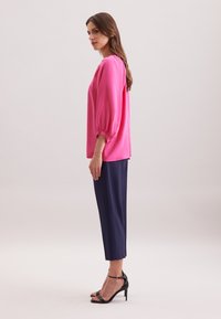 Blusa fucsia con maniche a sbuffo, abbinata a pantaloni navy a sigaretta e sandali neri con tacco alto. Tessuto liscio, minimalismo nei dettagli.