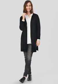 Czarny długi kardigan z otwartym przodem, noszony na białej koszuli z guzikami, w połączeniu z ciemnymi dżinsami i czarnymi butami, na neutralnym tle.