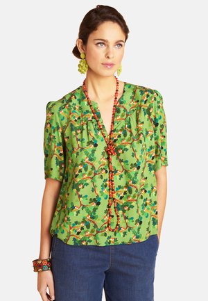 Frau trägt eine grüne Bluse mit Blumenmuster und Puffärmeln, eine rote Perlenkette, gelbe Ohrringe, ein gemustertes Armband und blaue Jeans.