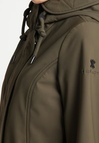 Olijfgroene jas met een capuchon, vervaardigd van textuurstof, met trekkoordsluiting en subtiele branding op de mouw.