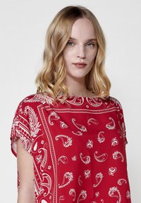 Blusa rossa con motivo paisley bianco, maniche corte e vestibilità ampia. Il tessuto sembra morbido con dettagli a bordi intricati.