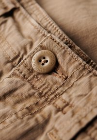 Tissu beige avec une finition texturée, présentant un unique bouton marron marqué "Superdry" et des coutures visibles sur la taille.