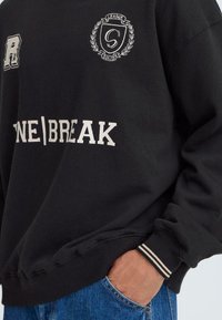 Mustas dress, millel on soonikkoesed, sisaldab valgeid graafikaelemente, sealhulgas kilbi logo ja teksti "ONE|BREAK" esiküljel.