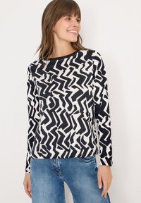 Langærmet top i sort og hvid med et abstrakt zigzag-mønster. Blødt stof med en rund halsudskæring. Kombineret med blå jeans.