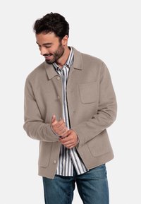 Veste en laine beige avec un col, fermeture boutonnée et une poche poitrine, portée sur une chemise rayée blanche et bleue, associée à un pantalon en denim.