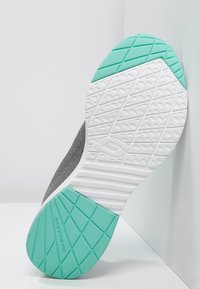 Skechers-sneakersula lutad mot vägg, med vit sula och strukturerade mönster samt gröna gummidetaljer vid häl och tå.