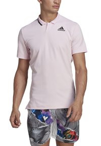 Polo shirt rose clair avec un col, présentant des accents noirs et le logo Adidas. Le tissu a une finition texturée.