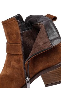Stivaletto in suede marrone con un accento in pelle nera, cerniera laterale e tacco impilato. L'interno presenta una fodera morbida e testurizzata.