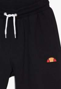 Pantalones de chándal negros de algodón con cinturilla elástica, cordón blanco y detalle de logo bordado en naranja y rojo en la parte inferior de la pierna izquierda.