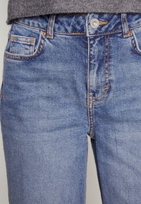 Lähikuva henkilöstä, joka käyttää sinisiä farkkuja, joissa näkyvät etutaskut, vyölenkit ja metallinen napitus harmaan neulevillapaidan alla.