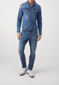 Ljusblå denimjacka med två bröstfickor och knappstängning, matchad med ljusblå jeans och vita sneakers med grå detaljer.