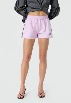 Femme portant un short de sport violet clair avec des rayures noires et des sandales noires à talons, debout, les mains derrière le dos.