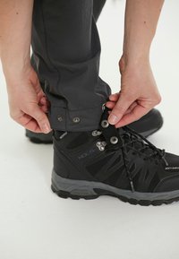 Zwarte waterdichte laarzen met textiel en rubberen zolen. Een persoon past de enkelboorden aan op grijze broek met drukknoopsluiting.