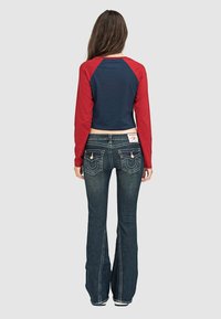 Rotes, langärmliges, verkürztes Oberteil mit navyfarbenem Körper. Kombiniert mit weit ausgestellten, dunkelblauen Jeans, die dekorative Rückentaschen und weiße Nähte haben.