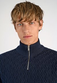 Marineblauwe ritssluitingtrui met een gestructureerd ruitpatroon en hoge kraag, voorzien van een zilveren rits en geribbelde details bij de kraag en zoom.
