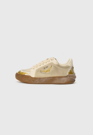 Beige sneakers made of suede and rubber. Features a curved white and gold pattern on the toe and side. Rounded toe and lace-up design.