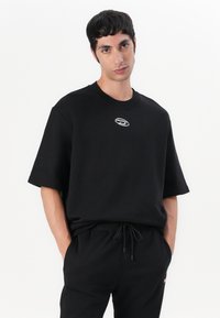 T-shirt en coton noir à manches courtes, col rond, avec un petit logo argenté sur la poitrine. Porté avec un pantalon de jogging noir assorti.