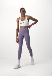 Balti sportinė liemenėlė su juodu „Nike“ logotipu, derinama su aukšto liemens purpurinėmis tampriomis kelnėmis, turinčiomis šonines kišenes. Modelis vilki baltus sportinius batelius ir kojines.