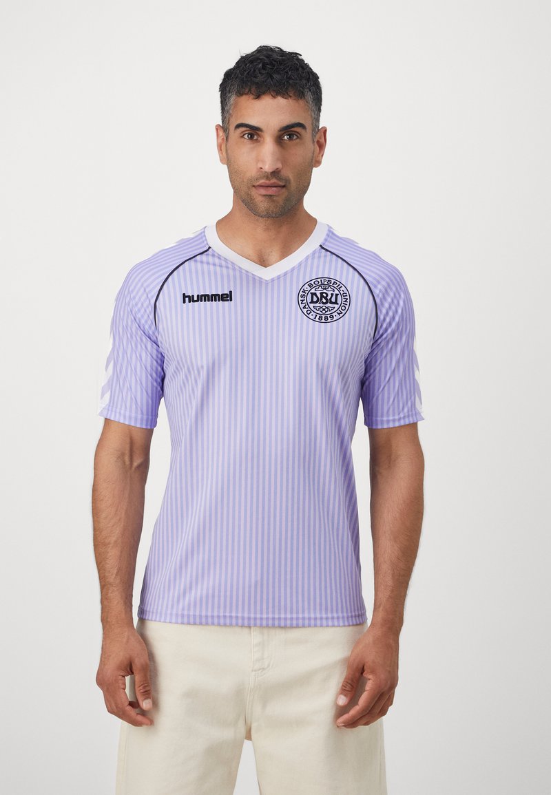 Uomo che indossa una maglia della squadra di calcio danese a righe viola chiaro con disegni a chevron bianchi sulle maniche e pantaloni beige, di fronte.