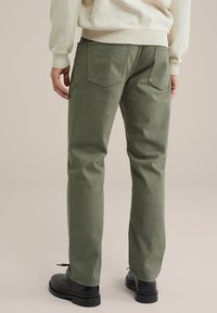 Olīvu zaļas kokvilnas bikses ar taisnu kāju dizainu, divām aizmugurējām kabatām un gludu tekstūru, apvienotas ar melniem ar auklēm sasienamiem apaviem.