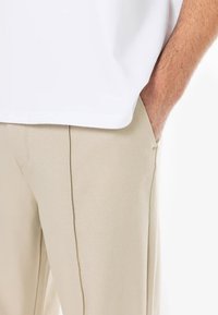 Pantalons beige sur mesure avec une texture lisse et des poches latérales, associés à un t-shirt blanc à la coupe large, montrant des lignes épurées et un design moderne.