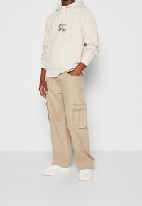 Beige cargobroek met zijzakken, gecombineerd met een crèmekleurige hoodie met een zwart logo. Zachte texturen en een ontspannen pasvorm. Witte sneakers maken de look af.