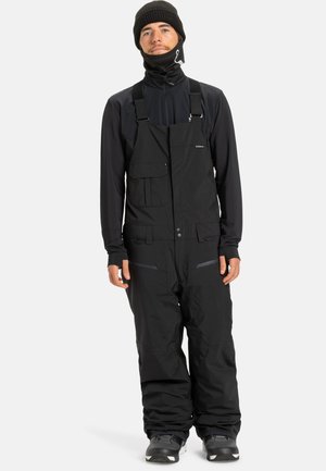 Homme portant une combinaison d'hiver noire, un t-shirt à manches longues noir, un bonnet noir, un masque facial et des bottes de neige, se tenant devant un fond uni.