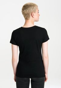 Person with short blonde hair wearing a plain black t-shirt and dark jeans, facing away against a white background.